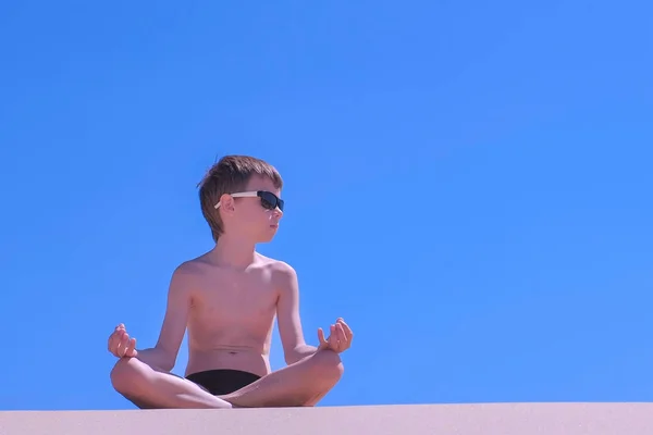 Niño en gafas de sol está meditando en pose de loto en la playa en el fondo del cielo . — Foto de Stock