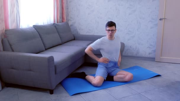 Engraçado nerd homem está fazendo torso torções exercício sentado no tapete em casa. Conceito de humor esportivo . — Vídeo de Stock