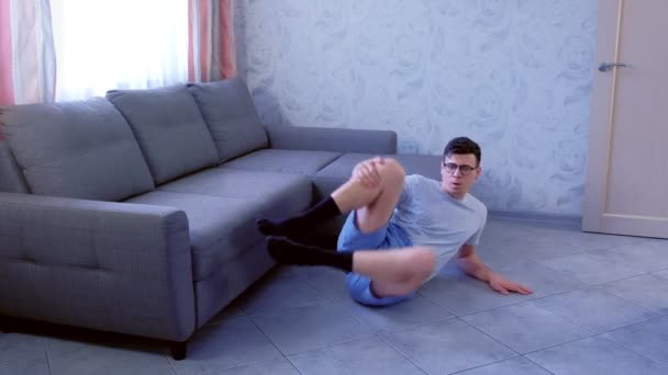 Homem nerd engraçado está fazendo exercícios de modelagem para as pernas que jazem no chão em casa perto do sofá. Conceito de humor esportivo . — Vídeo de Stock