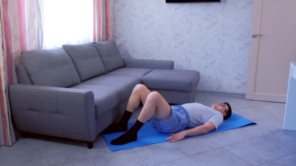 Funny nerd man is lifting legs lying on mat at home. Sport humor concept. — Stock Video