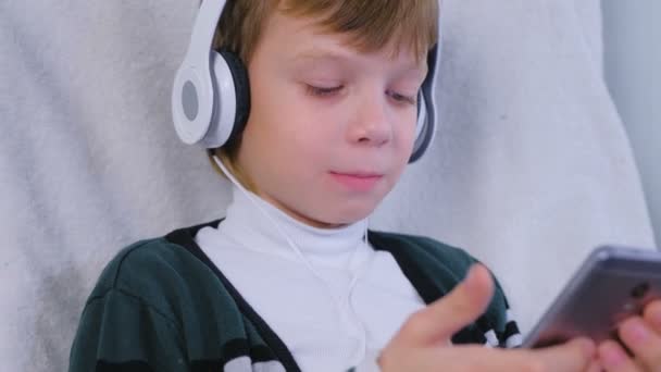 Menino está ouvindo música em fones de ouvido no smartphone e cantando sentado em poltrona em casa . — Vídeo de Stock