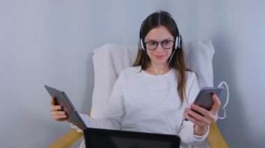 Kadın gözlük ve kulaklık koltukta oturan dizüstü bilgisayarda çalışıyor. Cep telefonu ve tablet elinde.