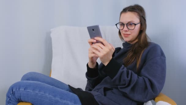 Young brunette woman in glasses types a message on a mobile phone sitting in armchair. — Stock Video