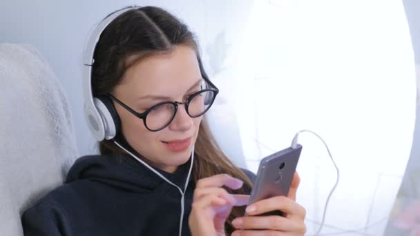 Жінка слухає музику в навушниках на смартфоні, сидячи в кріслі вдома . — стокове відео
