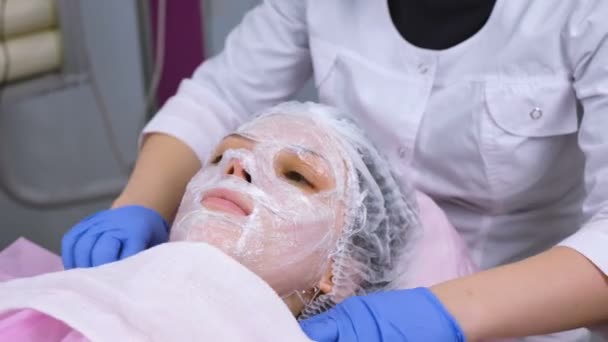 Kozmetikus eltávolítja a törülközőt Womans arcát. Cosmetologist eltávolítja a műanyag fólia a hidratáló maszk a Womans arca. — Stock videók