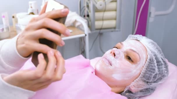 La mujer lee algo en un teléfono móvil tumbado en el procedimiento en la oficina de cosmetoloistas con una máscara en la cara . — Vídeo de stock