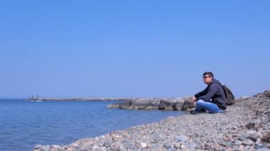 Adam gezgin güneşli bahar gününde deniz manzarası hayran deniz taş plajda oturuyor.
