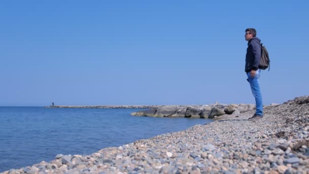 Homme voyageur avec sac à dos regarde la mer debout sur la plage de pierres de mer . — Video