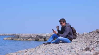 Man gezgin deniz plajında oturur akıllı telefon büyük haber almak hakkında mutlu.