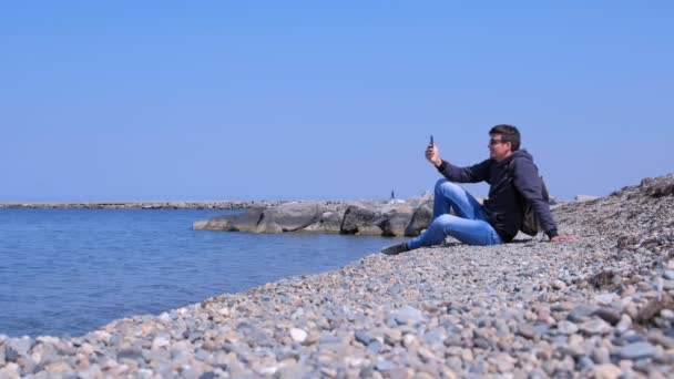 Guy Talks video chat in zijn smartphone zit op zee Stone Beach op vakantie. — Stockvideo