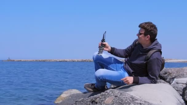 Homem fala vídeo chat telefone com pequeno tripé vem para praia do mar senta-se em pedra . — Vídeo de Stock