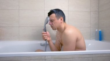 Yorgun adam küvette otururken yıkanıyor. Duştan yüzüne su dökmek.