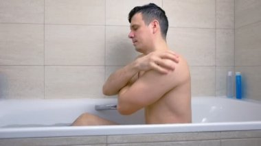 Adam küvette oturan duş jeli ile vücudunu yıkıyor.