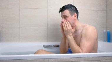 Adam küvette otururken yıkanıyor ve masaj hareketleri ile yüzüne scrub uygulayarak.