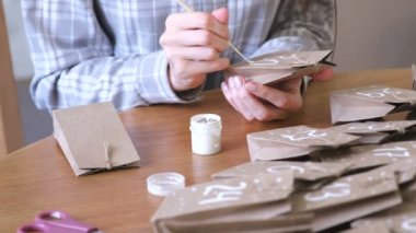 Kadın, çocuklar için Noel varış takvim yapar. Birkaç çanta üzerinde fırça ve beyaz boya ile yazar. Yakın çekim yakışıklı.