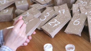 Kadın, çocuklar için Noel varış takvim yapar. Birkaç çanta üzerinde fırça ve beyaz boya ile yazar. Yakın çekim yakışıklı.