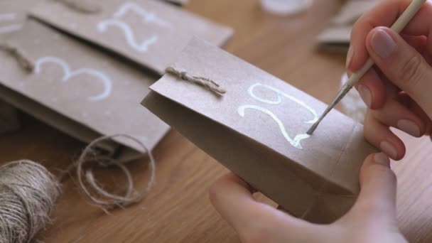 La mujer hace el calendario de Adviento de Navidad para los niños. Escribe un número en la bolsa por pincel y pintura blanca. Primer plano de las manos . — Vídeos de Stock