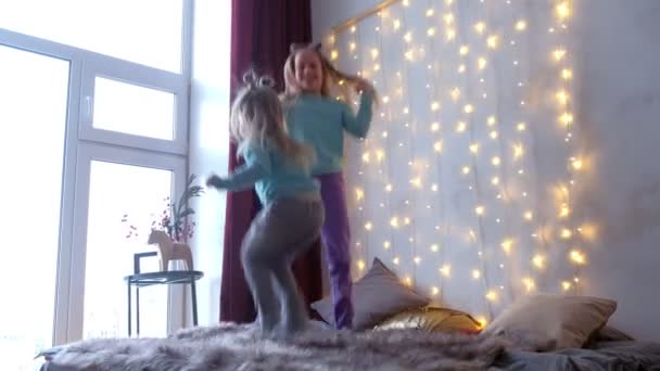 Twee meisjes zusters springen op bed thuis in kerst interieur met slingers. — Stockvideo