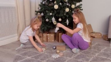 Noel ağacının yanında oturan oyuncak yıldızları ve topları birlikte oynayan iki kız kardeş.