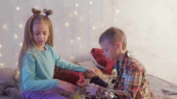 Chico y chica amigos jugando con guirnalda sentado en la cama en el interior de Navidad . — Vídeo de stock