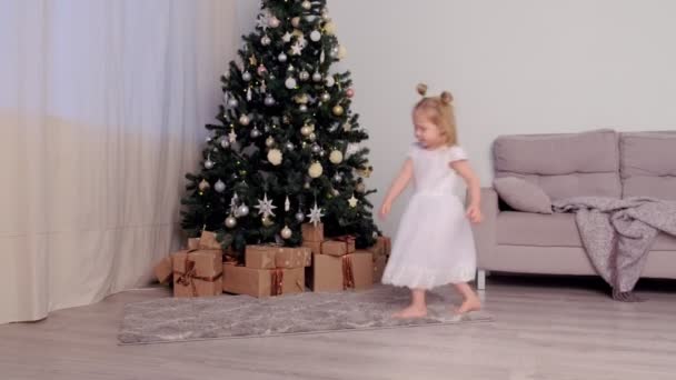 休日に自宅でクリスマスツリーの近くで踊る白いドレスの小さなかわいい女の子. — ストック動画