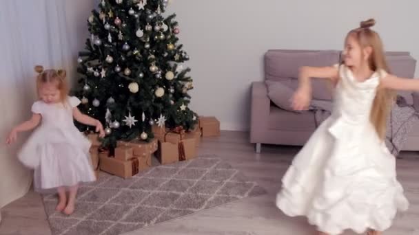 Dos hermanas en hermosos vestidos blancos giran y bailan cerca del árbol de Navidad . — Vídeos de Stock