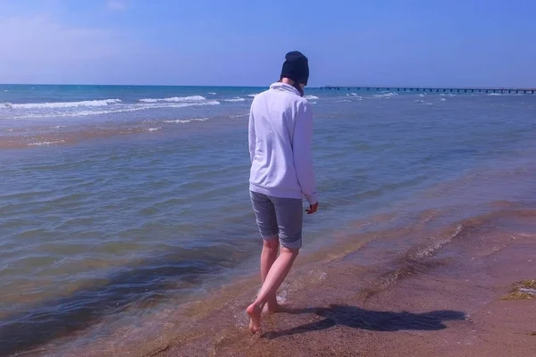 Mujer camina en el agua de mar en la playa de arena fuera de temporada lleva en sudadera con capucha y sombrero . —  Fotos de Stock