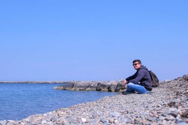 Adam gezgin güneşli bahar gününde deniz manzarası hayran deniz taş plajda oturuyor.