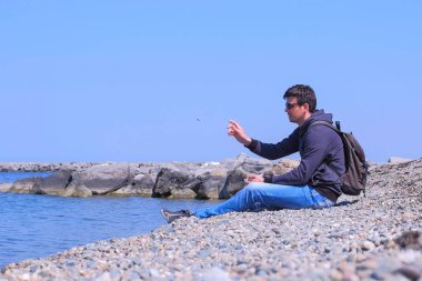 Adam turist gezgin taş plajda oturur ve suya taş atma.