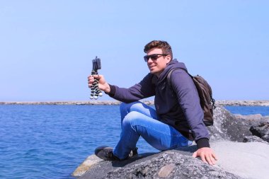 Küçük tripod ile Adam görüşmeler görüntülü sohbet telefonu deniz plajına gelir taş oturur.