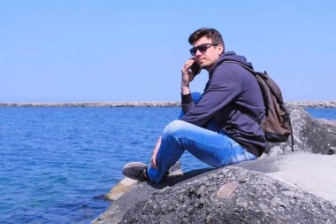 Adam gezgin akıllı telefon arıyor taş oturur deniz arka planda cevap bekler.