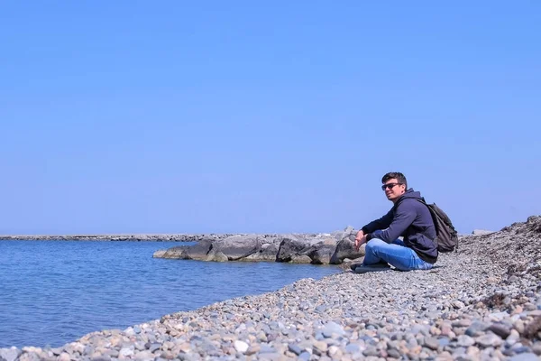 Adam gezgin güneşli bahar gününde deniz manzarası hayran deniz taş plajda oturuyor. — Stok fotoğraf