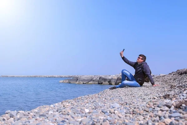 Adam gezgin deniz taş plajda oturuyor ve akıllı telefonda selfie yapıyor. — Stok fotoğraf