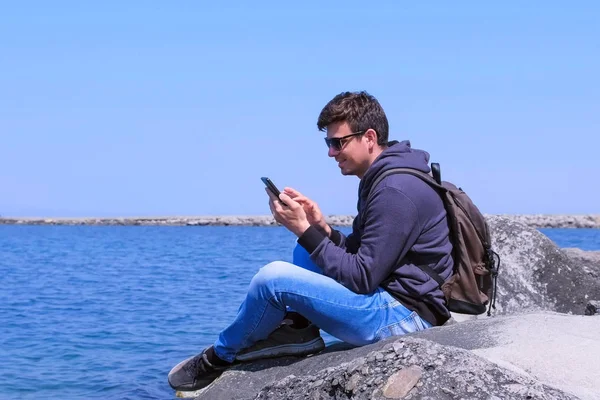 Hombre taveller chat tipo mensaje en el teléfono inteligente se sienta en piedra en la playa del mar . — Foto de Stock
