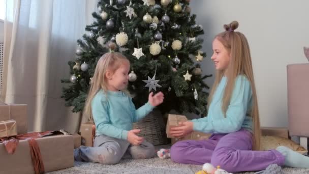 Κορίτσια αδελφές κουνώντας κουτί με το παρόν και παίζοντας καθισμένος κοντά χριστουγεννιάτικο δέντρο. — Αρχείο Βίντεο