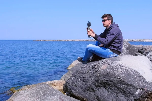 Man starts to record video on smartphone with tripod sits on huge stone at sea.