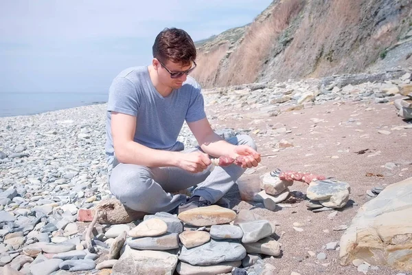 Människan förbereder shashlik sätta skivor av kött på spett sitter på havet sten strand med berg. — Stockfoto