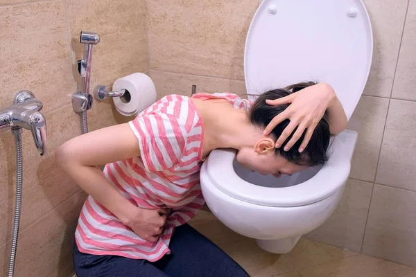 Mujer joven enferma con dolor de estómago está vomitando en el inodoro sentado en el suelo en casa, síntoma de gripe . —  Fotos de Stock