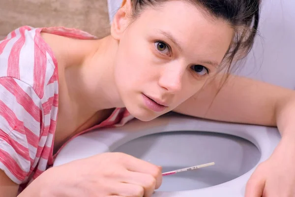Morgens Übelkeit. junge schwangere müde Frau mit Schwangerschaftstest in der Hand in der Toilette zu Hause und Blick in die Kamera. — Stockfoto