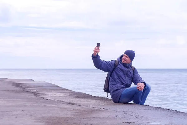 Man traveller makes selfie on smartphone sit on old waterfront at sea in winter. — Stock Photo, Image