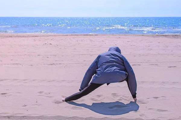 Vrouw in jas is warming-up op zee zandstrand stretching benen, achteraanzicht. — Stockfoto