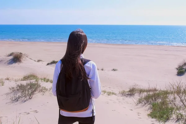 Girl travel stands on sand beach among dunes and look at sea vacation back view.