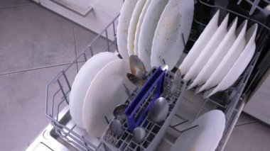 Mutfakta kirli beyaz yemekler ve çatal-bıçak takımı ile bulaşık makinesi sepeti.