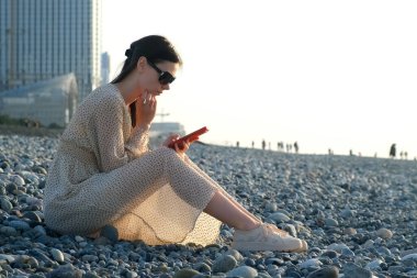 Batumi şehri Georgia 'da çakıl taşlı deniz kıyısında oturan akıllı telefona göz atan düşünceli bir kadın. Sosyal medyada telefonda kadın sohbeti. Yaz tatili, plajda dinlenme deniz turizmi..