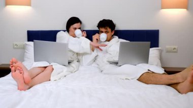 Kadın ve erkek dizüstü bilgisayarlarda projeleri tartışıyor ve çay içiyorlar. Yatakta beyaz bornozlarla yatıyorlar. Serbest meslek, mesafe, uzak iş kavramı, iş ve seyahat kavramı.