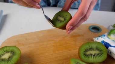 Kadın elleri, ev mutfağında ahşap tahtada kaşık kullanarak soyulmaktan kalan kivi parçalarını temizliyor. Yemek veya içecek için gerekli malzemeler. Yemek tarifi aşçısı. Meyve tatlısı hazırla..