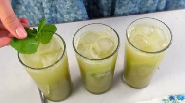 Evdeki mutfakta yeşil kivili limonatayı nane yapraklarıyla süsleyen bir kadın. Yemek tarifi, profesyonel aşçılık. Lezzetli meyve tatlısı hazırla. Ev yapımı içecek..