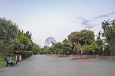 Bakü, Azerbaycan - 16 Mayıs 2025: Bakü, Azerbaycan 'daki Hazar Denizi kıyısındaki sahil parkı. Yeşil ağaçlar, yürüyüş yolu ve uzak bir Hilal Çarşısı manzarası..
