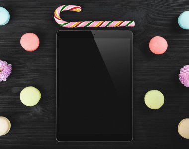 Siyah ahşap zemin üzerine üstten görünüm macarons, lolipop, dijital tablet ve pembe çiçekler ile tatil kavramının. Düz yatıyordu
