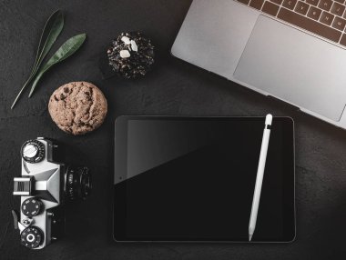 Dizüstü bilgisayar, dijital tablet ve fotoğraf makinesi, siyah bir masaüstü üstten görünüm. Kahve molası tatlılar, üstten görünüm
